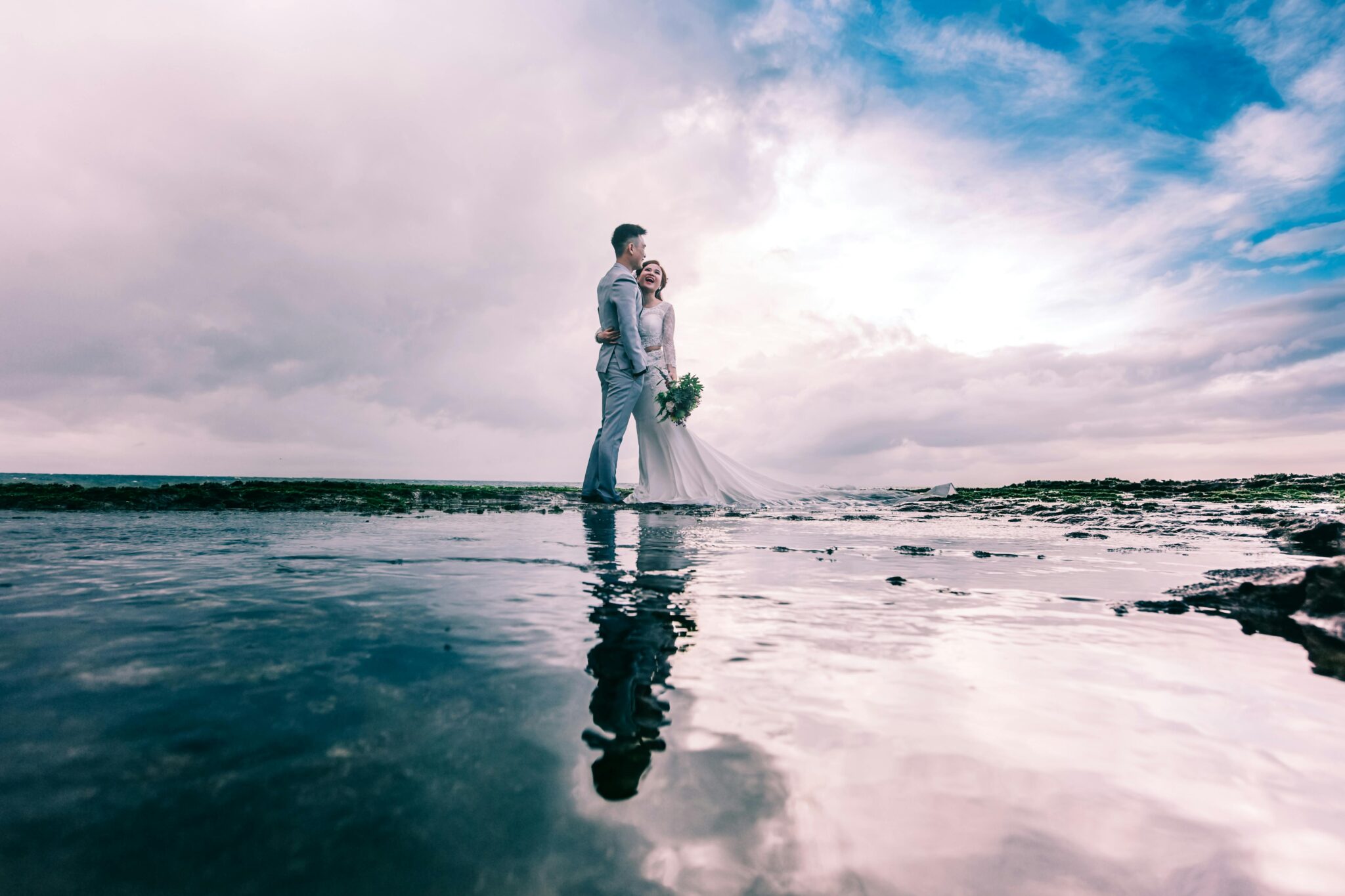 A joyful couple embracing on the beach in wedding attire under a dramatic sky.